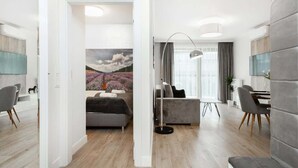 A modern living room with a grey sofa, a dining area with a table and chairs, and a large window with curtains. - Modern Apartment with Air Conditioning | 2 Bedrooms | Parking (Warszawa)