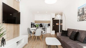 A modern living room with a flat-screen TV, a dining area with a table and chairs, a grey sofa, and a kitchen in the background. - Modern Apartment with Air Conditioning | 2 Bedrooms | Parking (Warszawa)