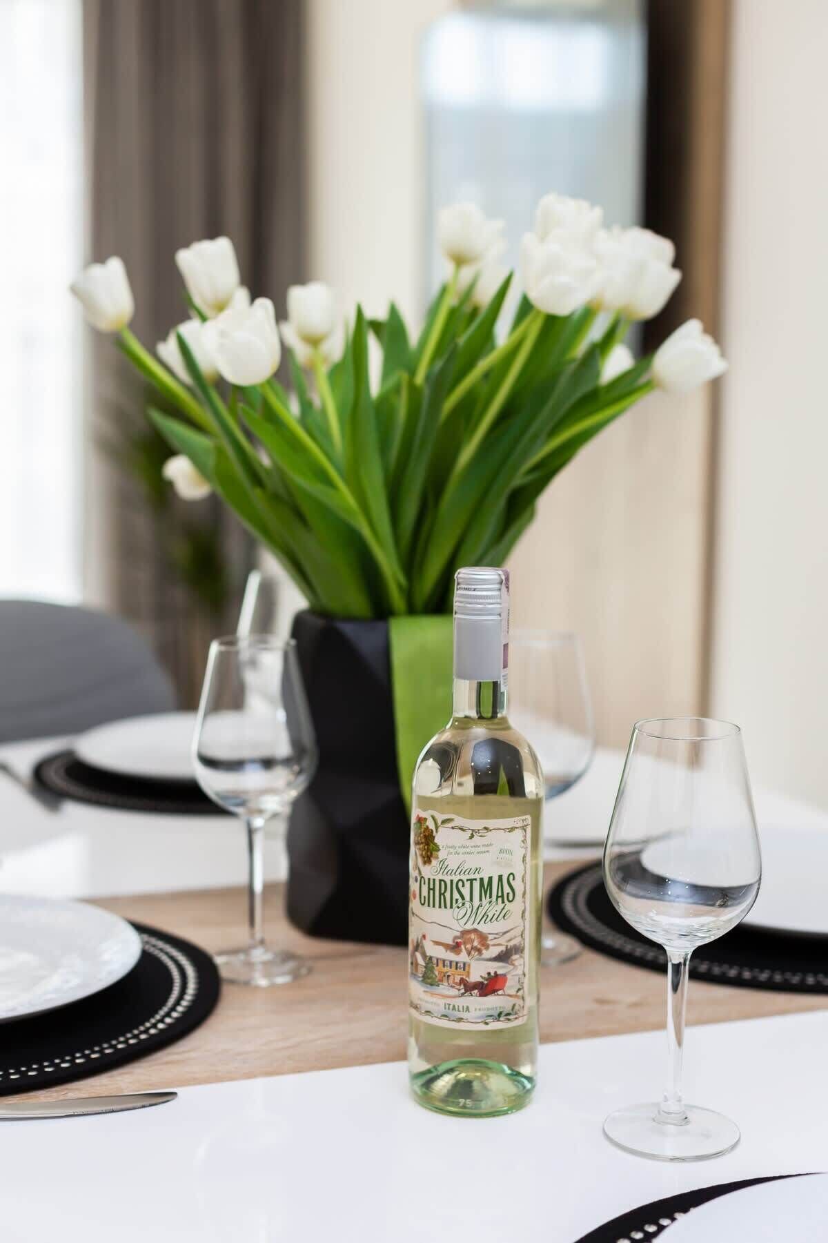 En flaske italiensk hvid julevin, to tomme vinglas og en vase med hvide tulipaner på et bord.
