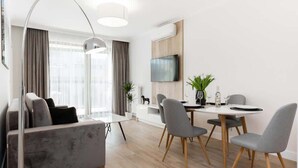 A modern living room with a grey sofa, a dining table with four chairs, a TV mounted on the wall, and a large window with curtains. - Modern Apartment with Air Conditioning | 2 Bedrooms | Parking (Warszawa)