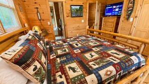 Une pièce de cabane en bois avec une couvre-lit matelassé, des murs en bois, un téléviseur et une porte donnant sur une autre pièce.