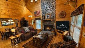 L’intérieur d’un chalet confortable, avec des murs en rondins de bois, une cheminée en pierre, un canapé en cuir, un coin repas et un téléviseur diffusant l’émission « Lord’s View ».