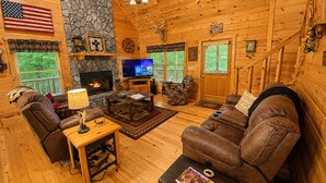 L’intérieur d’une cabane en rondins, avec une cheminée en pierre, un téléviseur, des canapés en cuir et un ventilateur de plafond.