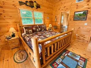 Un dormitorio de cabaña de madera con una cama, dos mesitas de noche, un ventanal, una puerta y una alfombra.