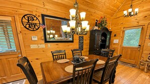 A wooden interior with a dining table, chairs, a chandelier, a fireplace, and a mirror.