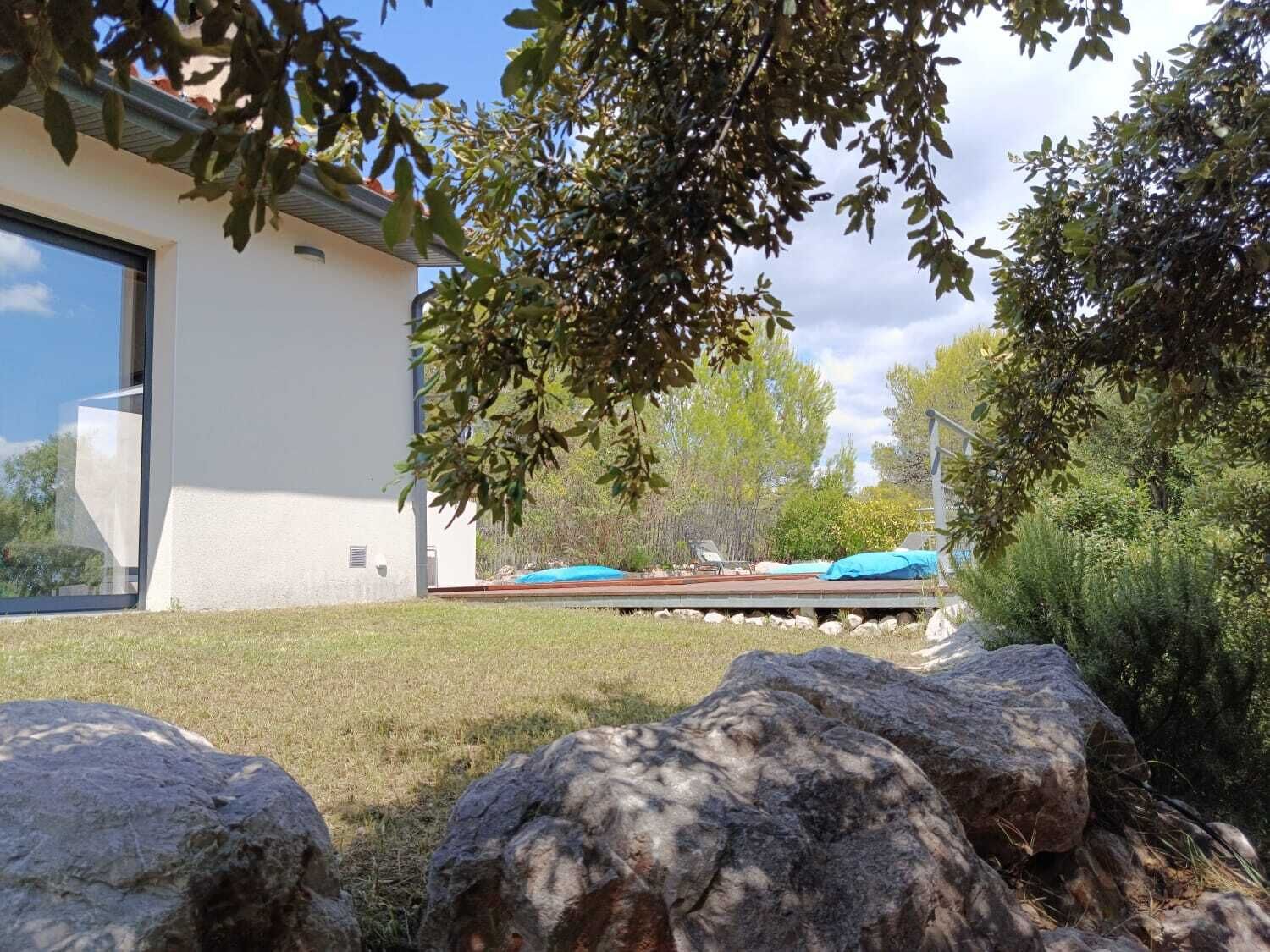 Un edificio moderno con un amplio ventanal, un patio con mobiliario exterior y un paisaje natural con rocas y árboles.