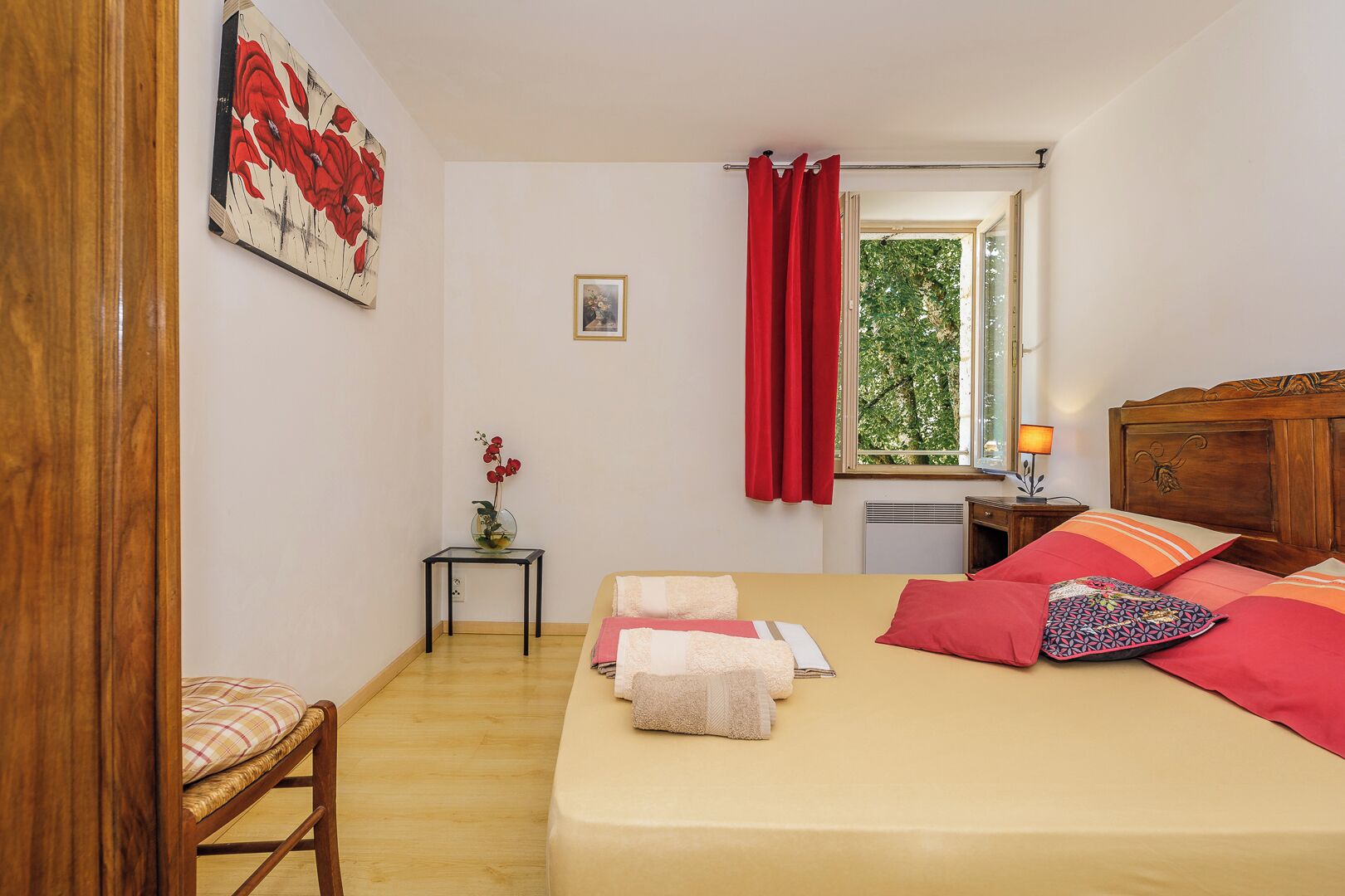 A bedroom with a bed, a nightstand, a window with red curtains, a small table with a vase of flowers, and a painting on the wall.