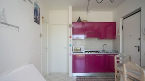 A compact living space with a kitchenette featuring pink cabinets, a microwave, and a stove. There is a white door, a small rug, and a wooden chair. - M247 - Marcelli, new two-room apartment with terrace and garage (Marcelli)