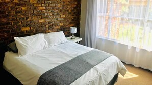 Een hotelkamer met een bakstenen muur, een bed met witte lakens, een nachtkastje met een lamp en een raam met uitzicht op groen.