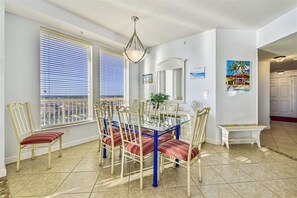 A dining area with a glass table and chairs, a hanging light fixture, and a view of the ocean through large windows. - Stunning West End View, Spring/Summer open, book your getaway now! (Panama City Beach)