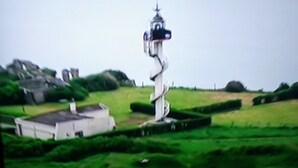 Un phare doté d’un escalier en colimaçon, entouré d’une pelouse verdoyante et avec quelques bâtiments visibles au loin.