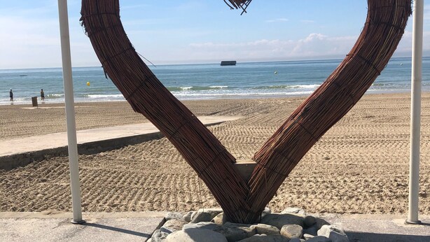 Une sculpture en forme de c œur, réalisée à partir d’un matériau tissé, est posée sur un socle en béton agrémenté de rochers, et offre une vue sur une plage, un ciel dégagé et l’océan.
