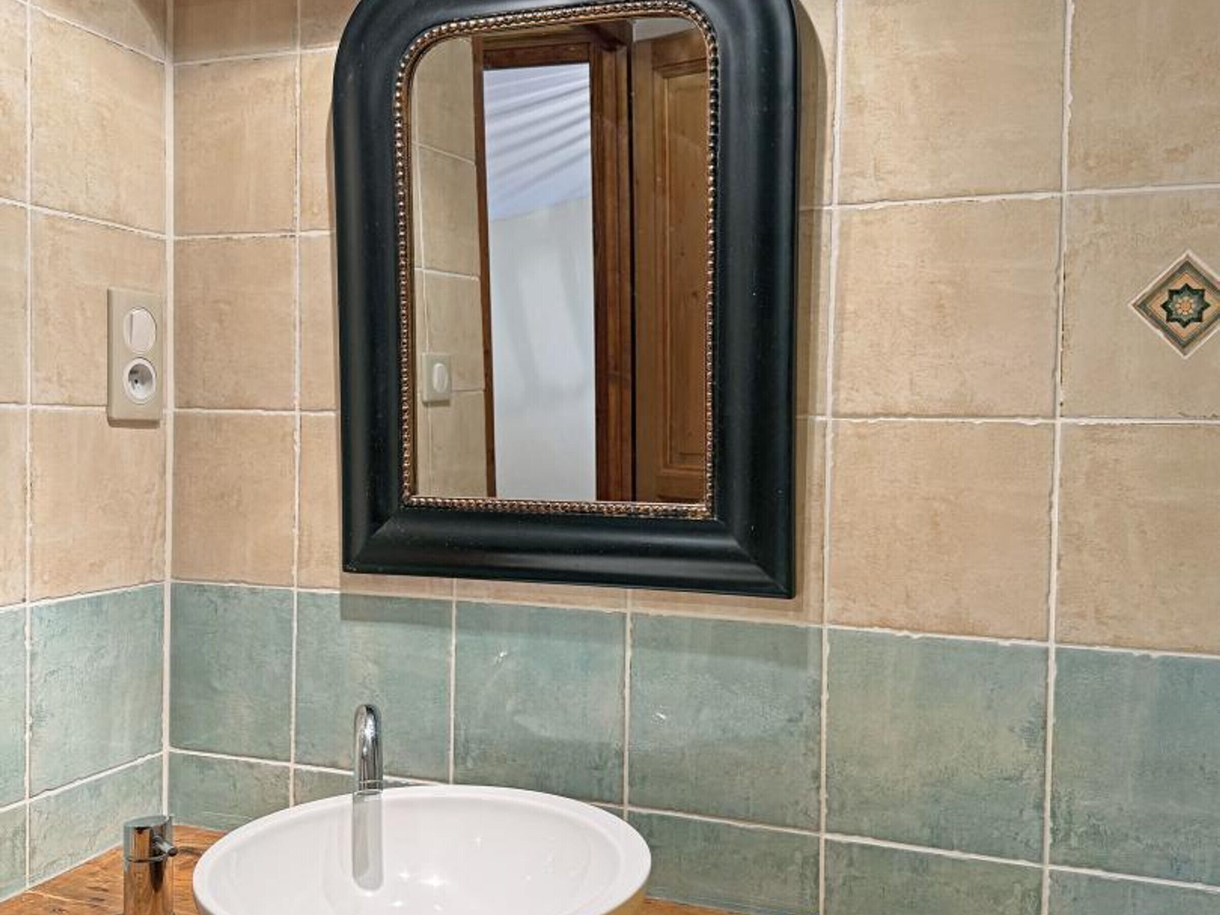 Ein Badezimmer mit einem weißen Waschbecken, einem Spiegel mit schwarzem Rahmen sowie beige- und turquoise farbenen Fliesen.