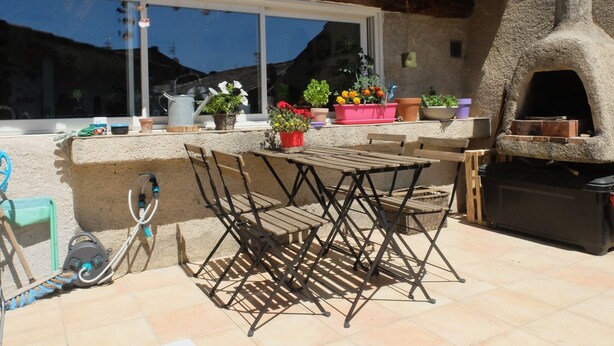 An outdoor patio with a table and chairs, a window with potted plants, and a stone oven. - Charming village house in the Luberon (Cadenet)