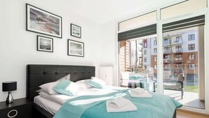 A modern hotel room with a large bed, a nightstand, a lamp, and a balcony with a view of other buildings.