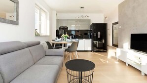 A modern living room with a grey sofa, a dining table with chairs, a kitchen area with a fridge, and a TV on a stand.