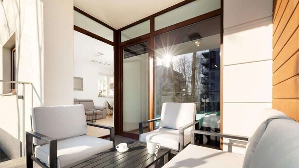 A modern outdoor seating area with white chairs and a wooden table, large glass windows, and a view of a building across the street.