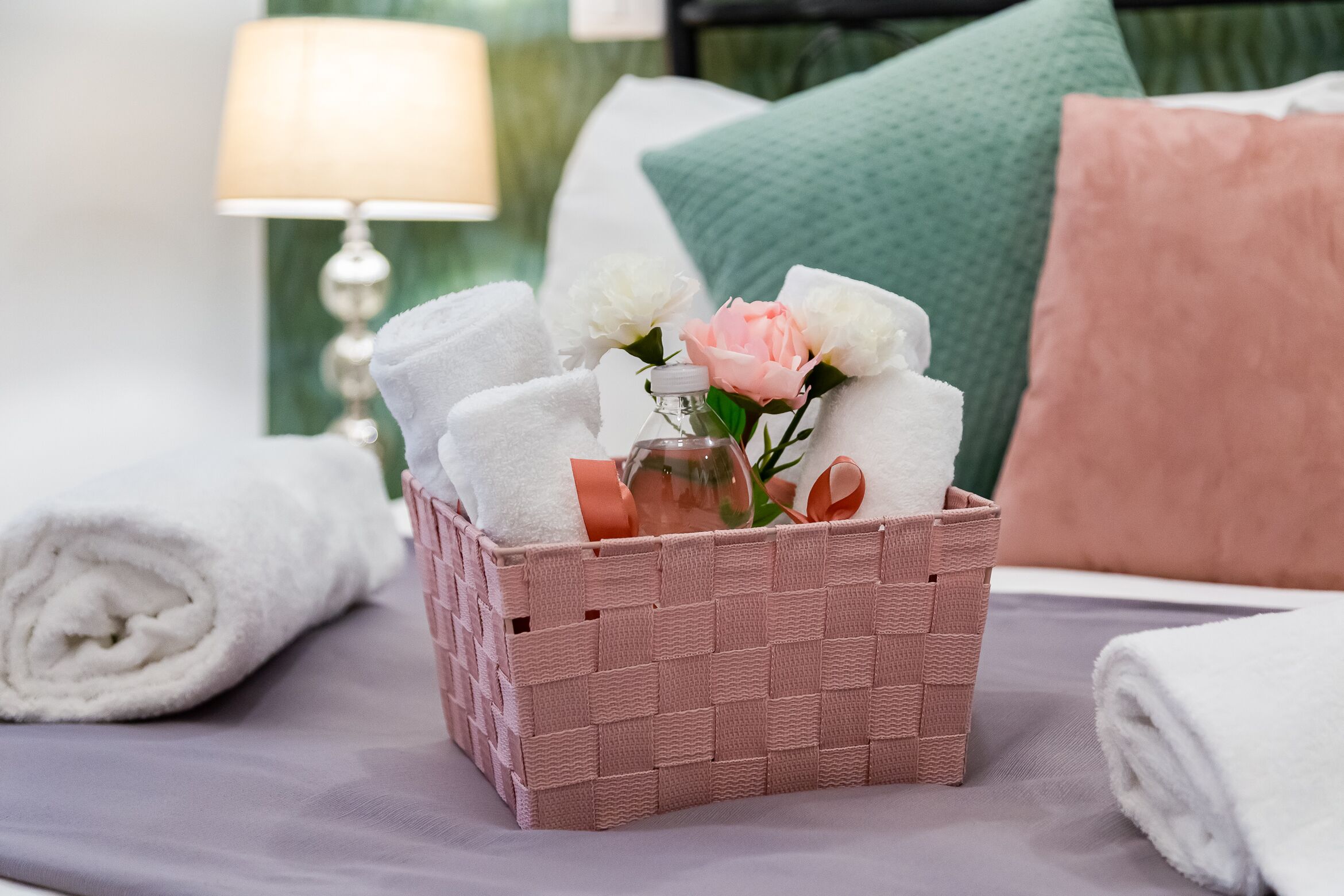 Un cesto di vimini pieno di asciugamani bianchi arrotolati, un fiore rosa e una bottiglia di vetro su un letto con un cuscino verde e uno bianco sullo sfondo.