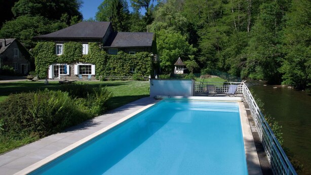 A swimming pool with a white deck, surrounded by lush greenery and a house with a dark roof. - Gite on the edge of the Arac in a green setting with swimming pool (Aleu)