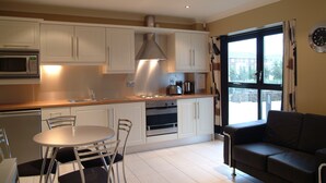 A compact kitchen with white cabinets, a microwave, and a stove. There is a dining table with chairs and a sofa in the living area. - Cuirt na Coiribe 4 bedroom Apartment (sleeps 5) (Galway)