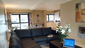 A modern living room with a sectional sofa, a coffee table, and a laptop on a side table. There is a dining area in the background with a table and chairs. - Cuirt na Coiribe 4 bedroom Apartment (sleeps 5) (Galway)