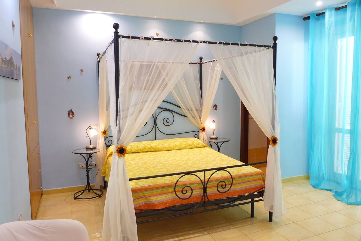 A four-poster bed with a canopy, a bedside table with a lamp, and a view of the outside through a window with blue curtains.