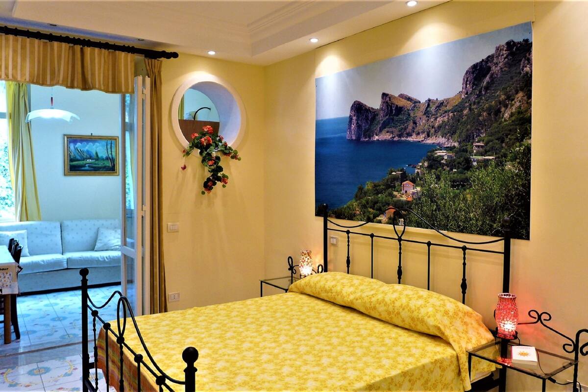A bedroom with a bed, a window with curtains, a painting of a coastal landscape, and a small table with a vase.
