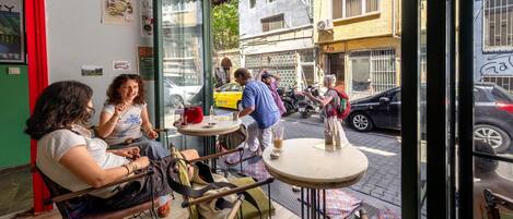 Um café ao ar livre com mesas e cadeiras, entrada por uma porta de vidro e vista para a rua.