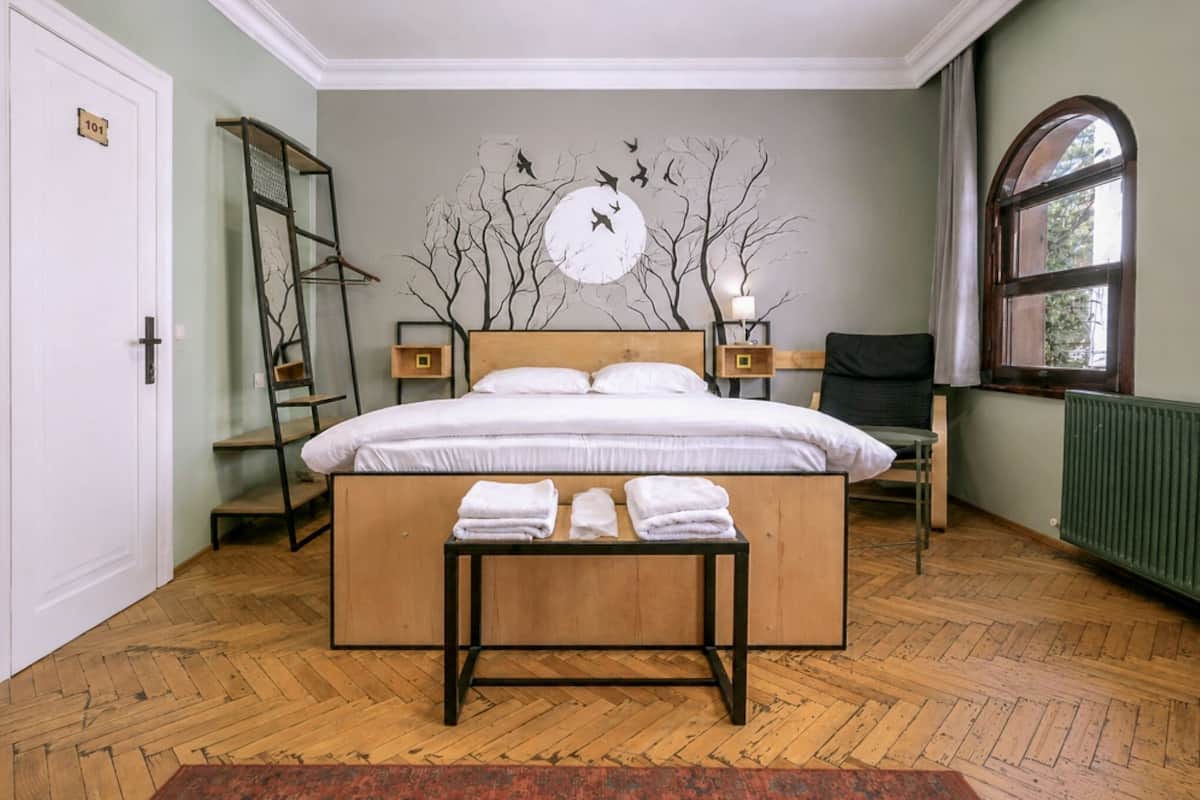 Un dormitorio con cama, mesita de noche, lámpara, silla, ventana y una obra de arte en la pared.