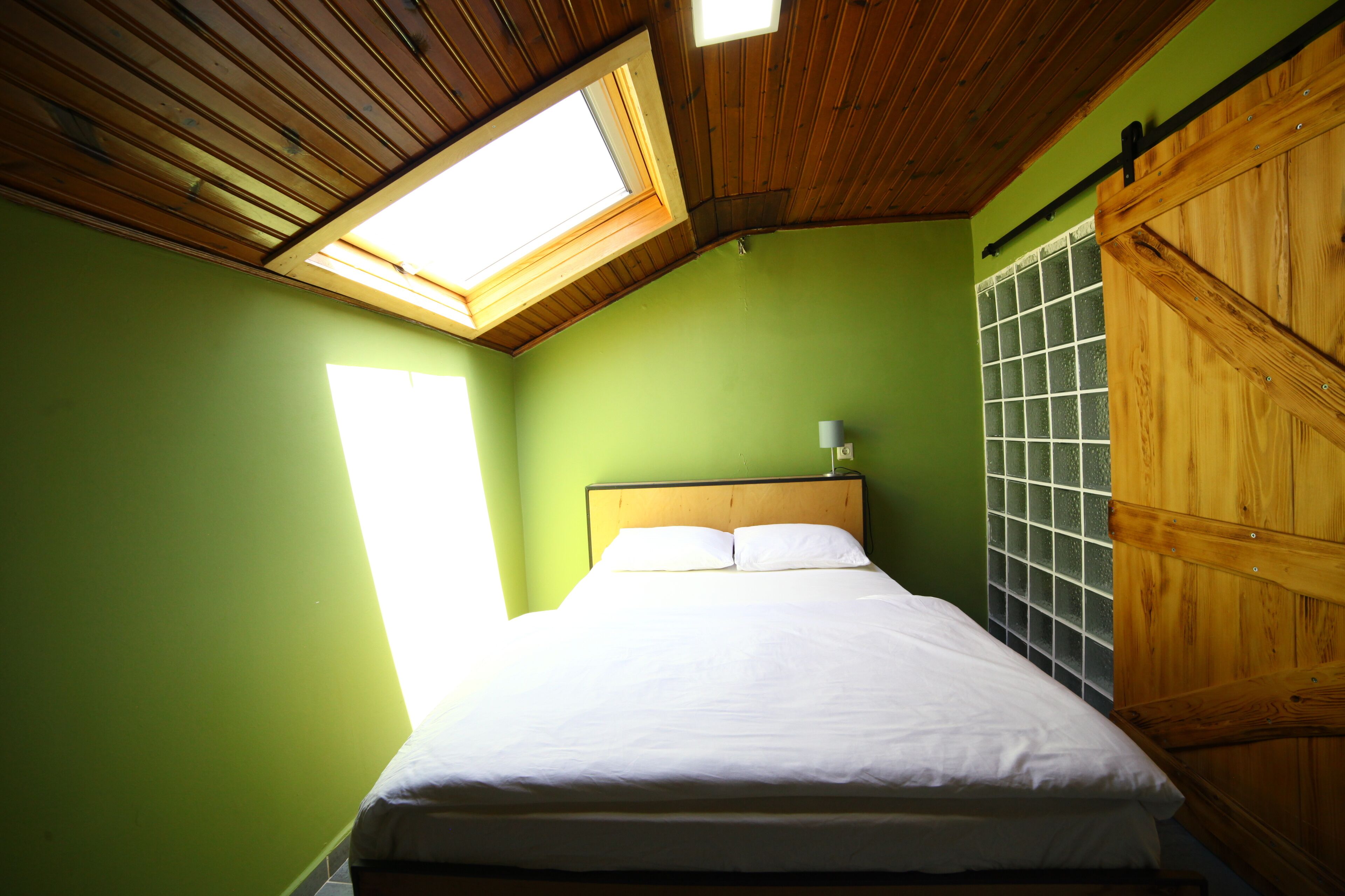 Un dormitorio con tragaluces, una cama con ropa blanca, cabecera de madera y pared verde.