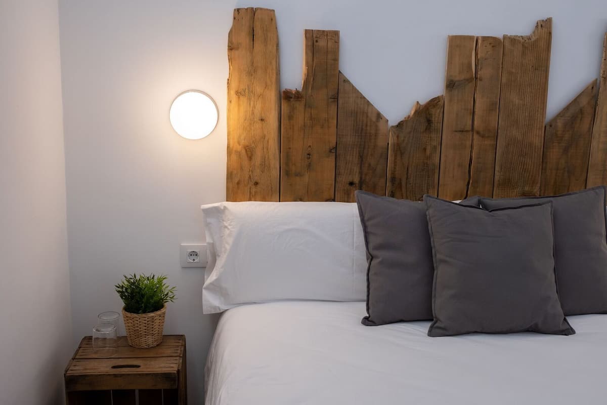 A bedroom with a wooden headboard, white bedding, and a bedside table with a plant and glasses.
