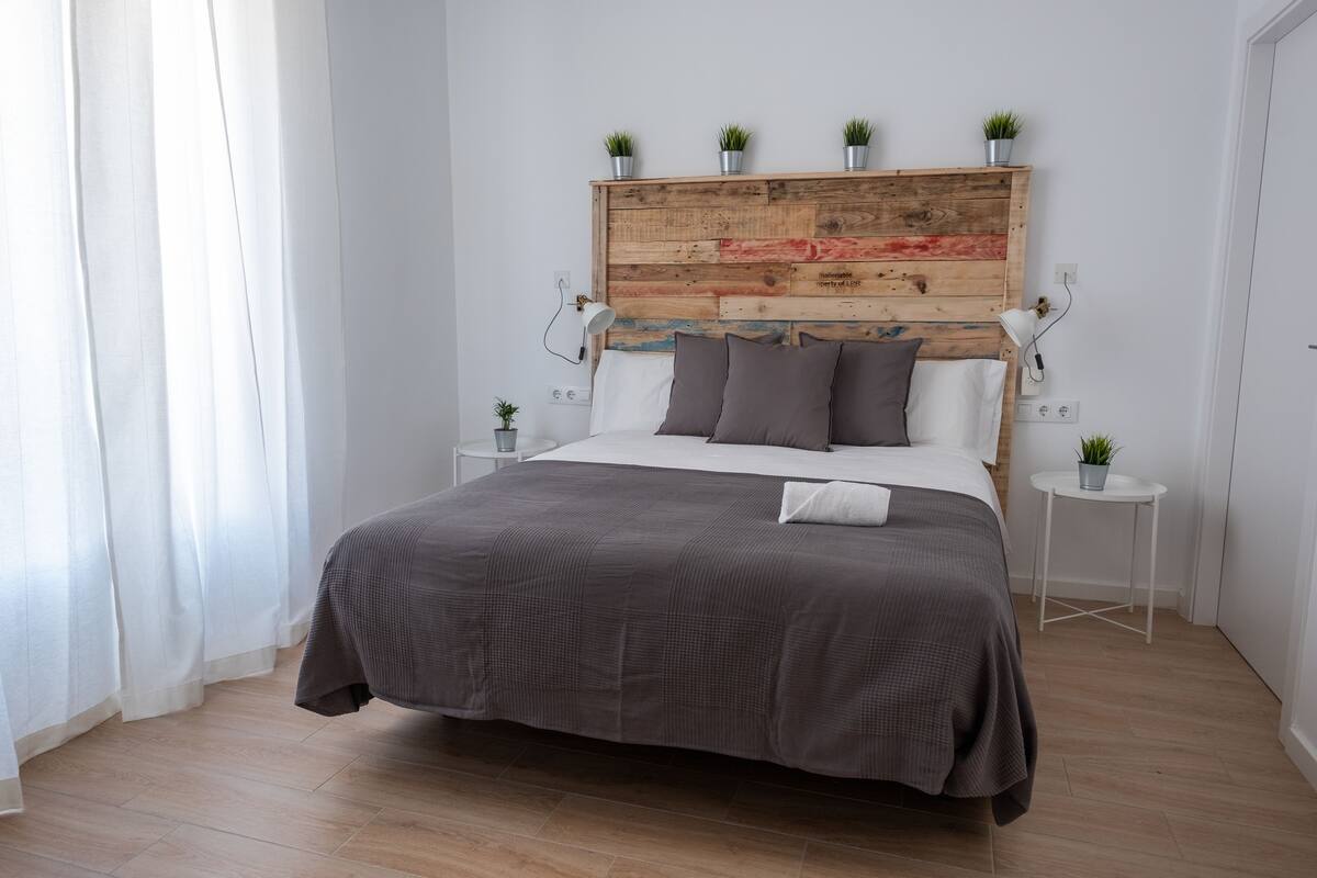 A bedroom with a wooden headboard, a bed with a gray bedspread, two bedside tables with lamps, and small potted plants.
