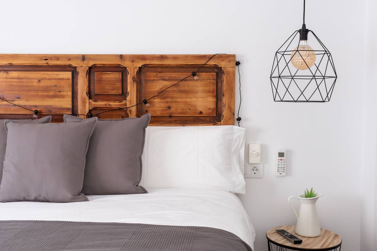 A bedroom with a wooden headboard, a bed with grey and white bedding, a small table with a plant and a remote control, and a geometric pendant light.