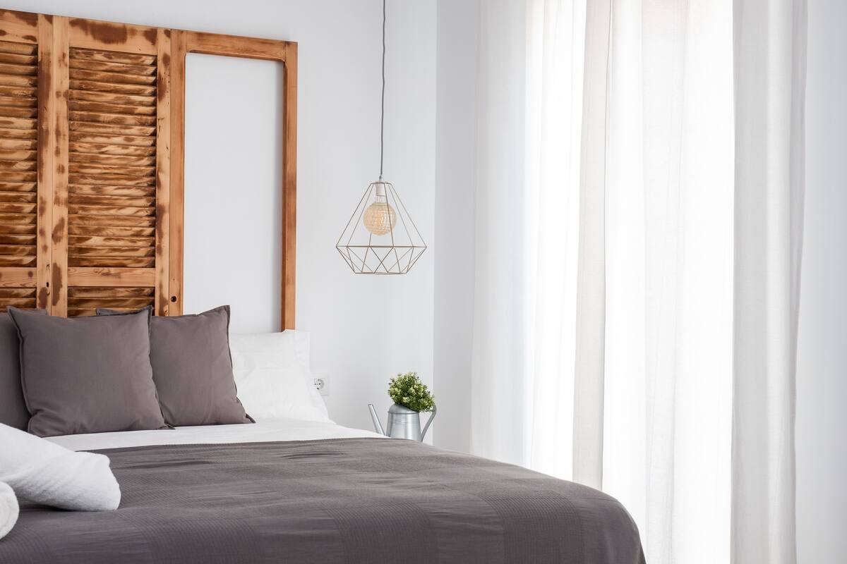 A modern bedroom with a wooden headboard, a geometric pendant light, a small plant, and a neatly made bed.