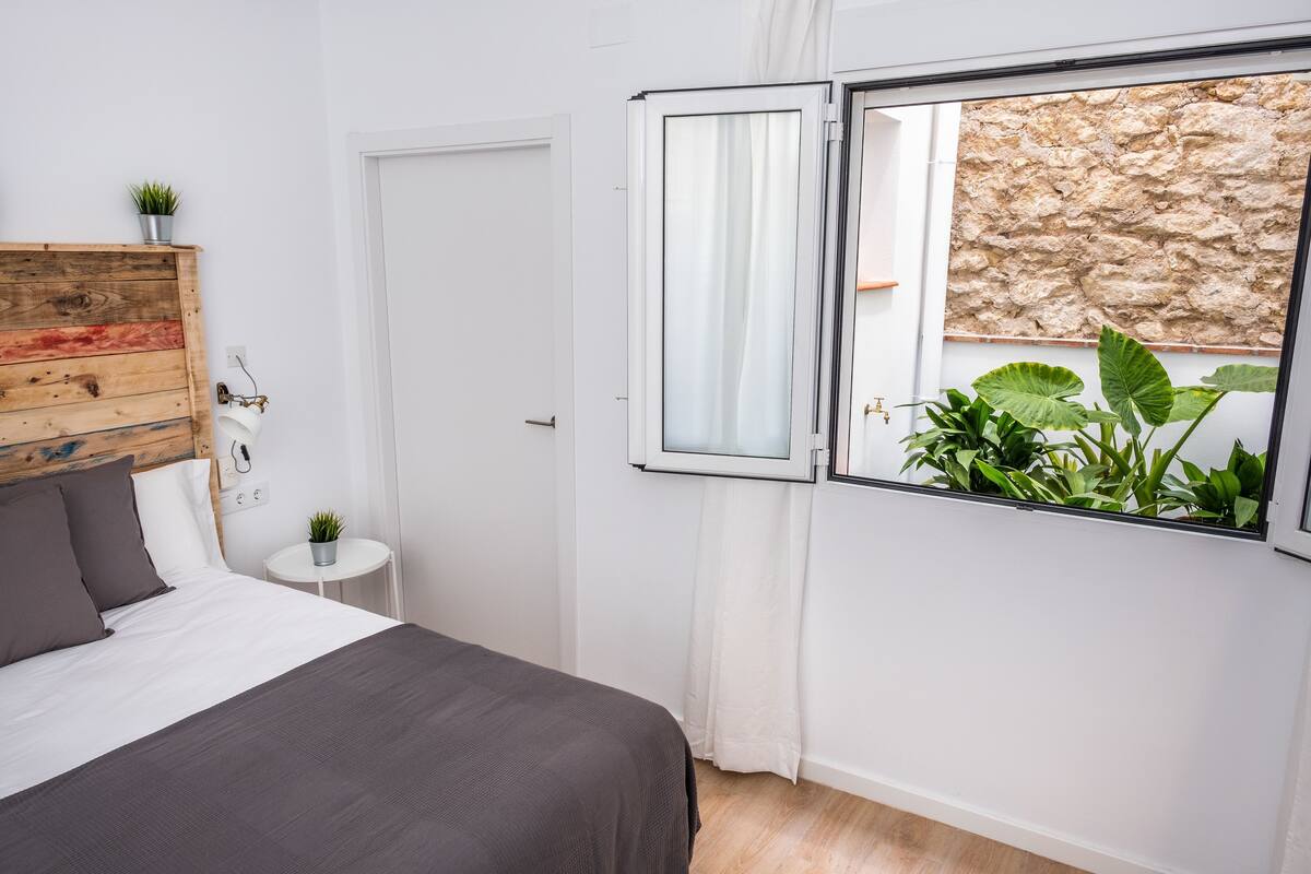 A bedroom with a bed, a small table with a plant, a wooden headboard, and a window with a view of a stone wall.