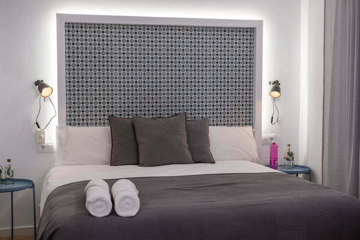 A neatly made bed with a grey and white patterned headboard, two bedside lamps, and a small table with a pink bottle and a glass.