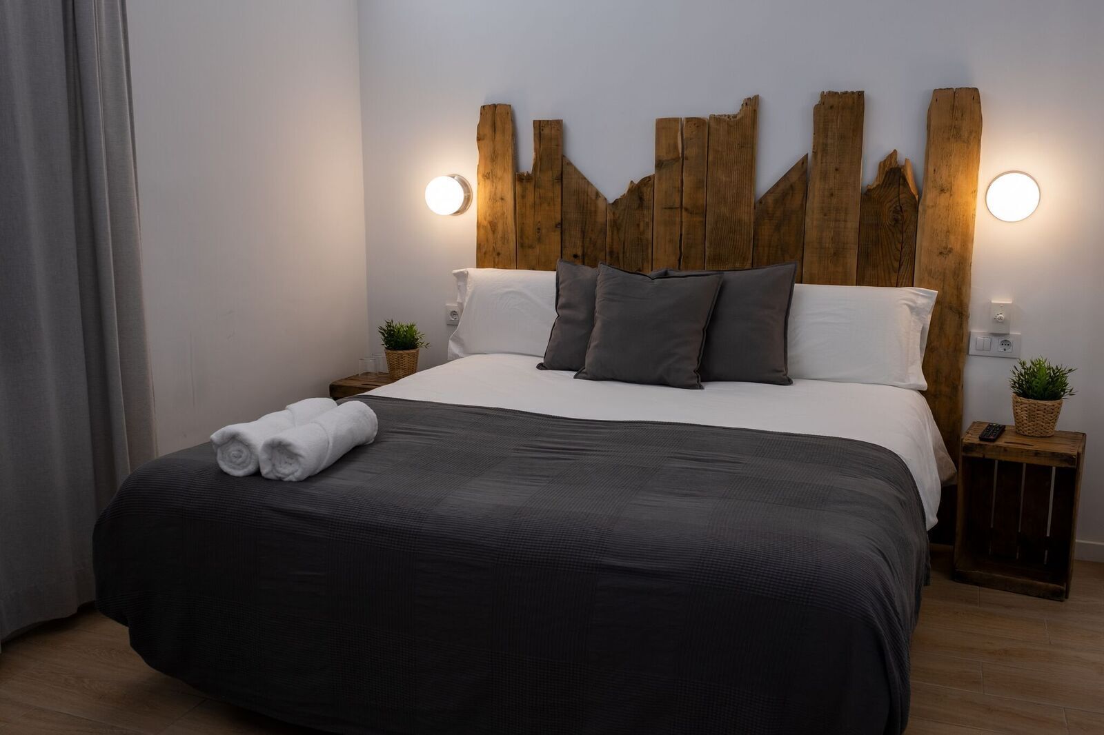 A neatly made bed with a dark grey comforter and pillows, a wooden headboard, bedside tables with lamps, and a potted plant on one table.