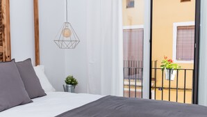 Un dormitorio con cama, una lámpara colgante, una pequeña planta en maceta y vistas a un balcón con barandilla y un edificio al fondo.