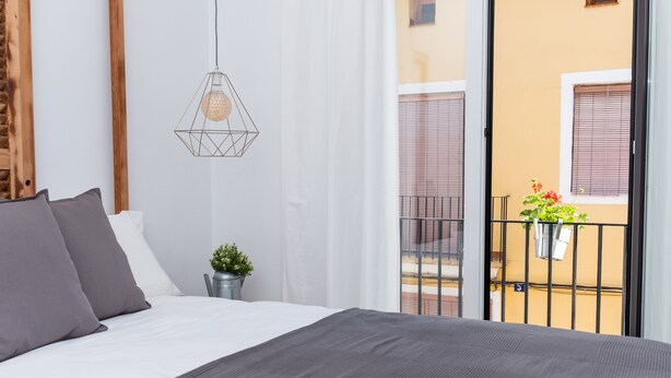 Une chambre avec un lit, un luminaire suspendu, une petite plante en pot et une vue sur un balcon avec une balustrade et un immeuble en arrière-plan.