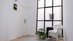 Una habitación con un ventanal grande, una silla de mimbre blanca, una mesita con libros y una planta, y una puerta con el número.