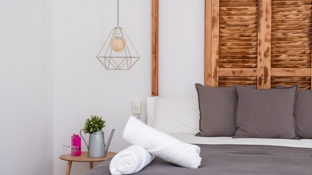 Una cama bien hecha con una colcha gris, una toalla blanca enrollada y dos almohadas grises. Una lámpara colgante de diseño ilumina la cama, y se ve una pequeña mesita de noche con una botella de agua rosa, un vaso transparente y una planta en maceta.