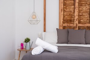 A neatly made bed with a grey bedspread, a rolled white towel, and two grey pillows. A geometric pendant light hangs above the bed, and a small wooden side table with a pink water bottle, a clear glass, and a potted plant is visible. - Gómez Rooms (Xàtiva)