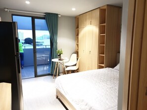 A hotel room with a bed, a desk, a chair, a wardrobe, and a sliding glass door leading to a balcony.