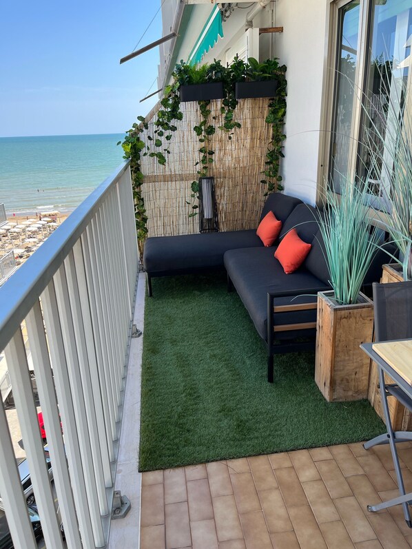 A balcony with a black sofa, red cushions, and a wooden planter with tall green plants. - Appartement DreamAppart Lido di Jesolo Venise (Lido di Jesolo)