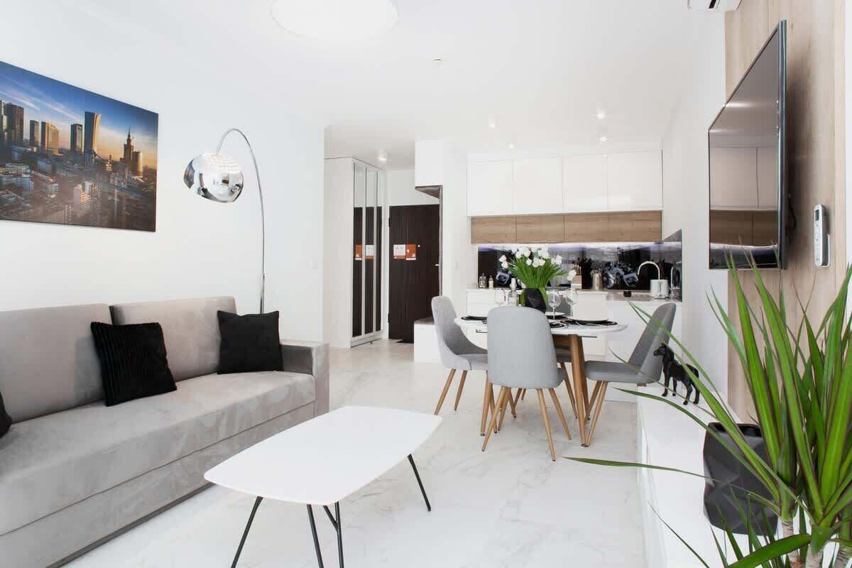 A modern living room with a sofa, a dining table with chairs, a flat-screen TV mounted on the wall, and a cityscape painting.