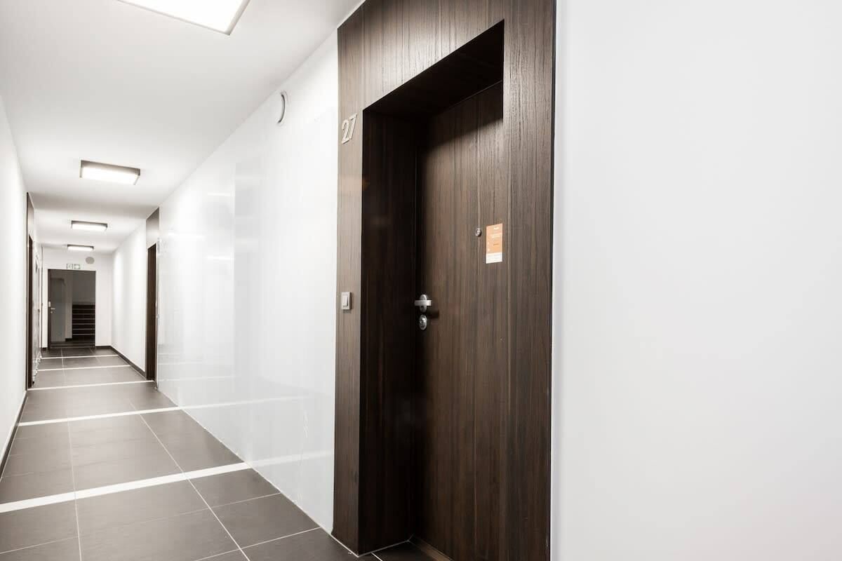 Un largo pasillo estrecho con una puerta de madera oscura a la derecha, paredes blancas y suelo de baldosas.