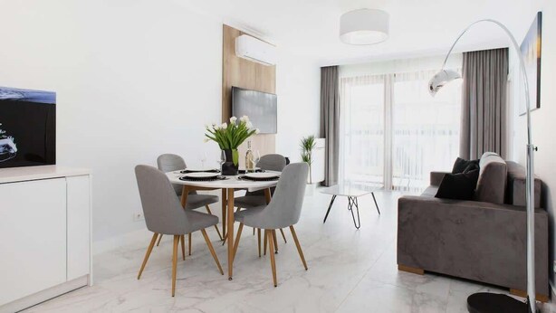A modern living room with a grey sofa, a dining table with chairs, a TV, and a large window with curtains. - Air-conditioned Apartment with Balcony | Parking and Close to the Metro (Warszawa)