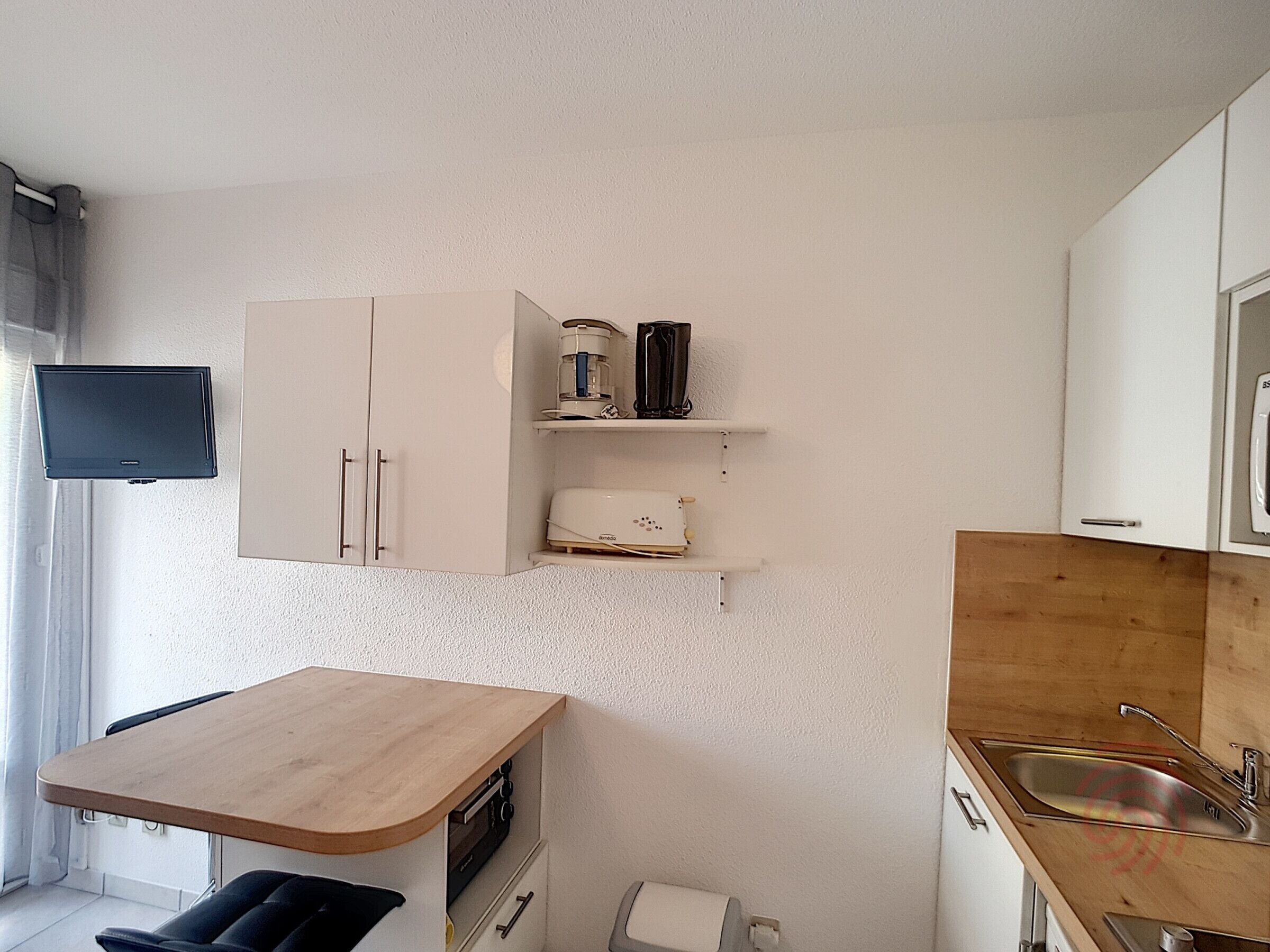 Une cuisine compacte avec des meubles blancs, un plan de travail en bois, un four encastré, un évier et un téléviseur à écran plat fixé au mur.