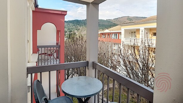 Un balcón con una mesa y sillas, una puerta roja y vistas a edificios y colinas.