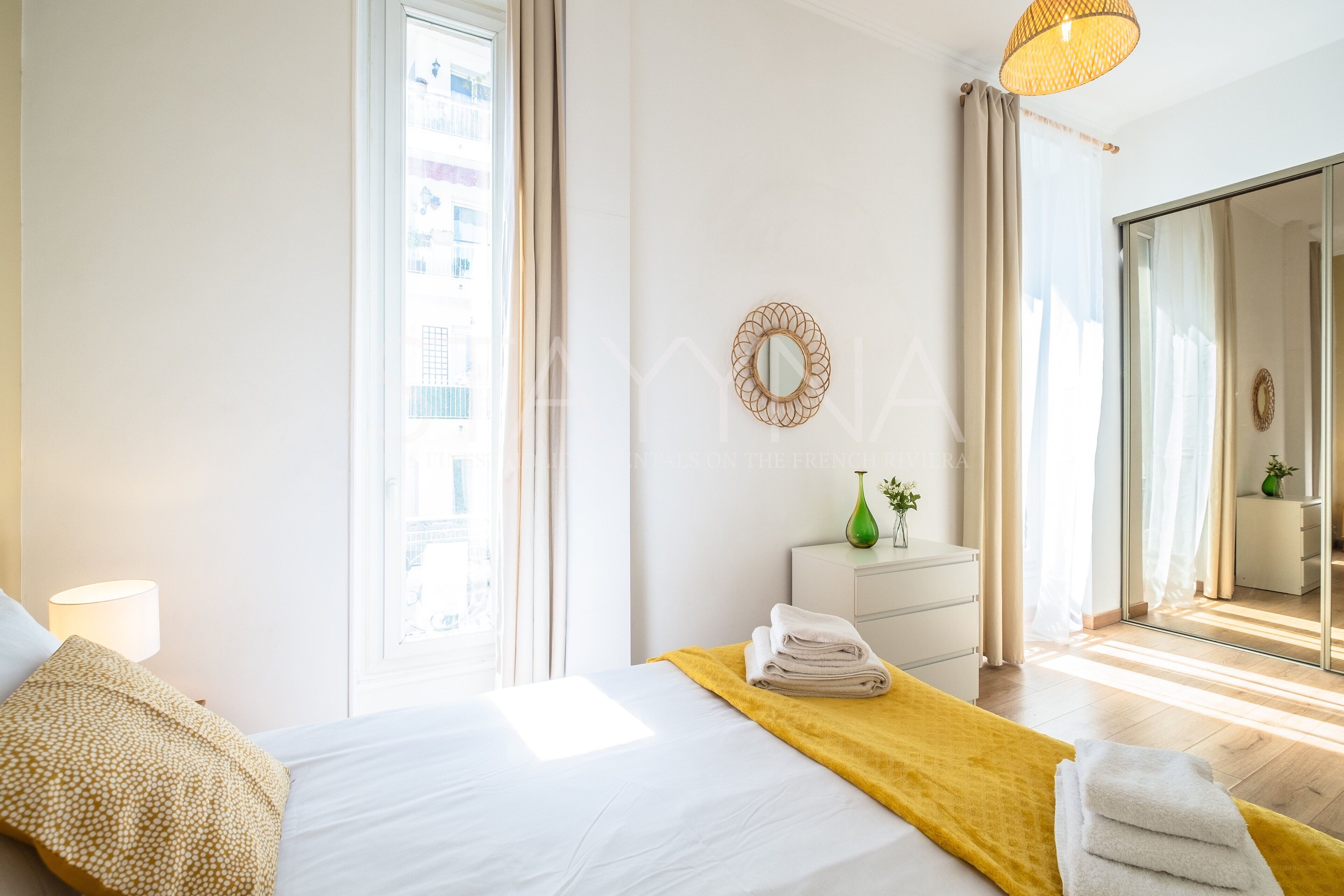 Ein Hotelzimmer mit Bett, Nachttischlampe, Kommode, Spiegel und einem Fenster mit Vorhängen.